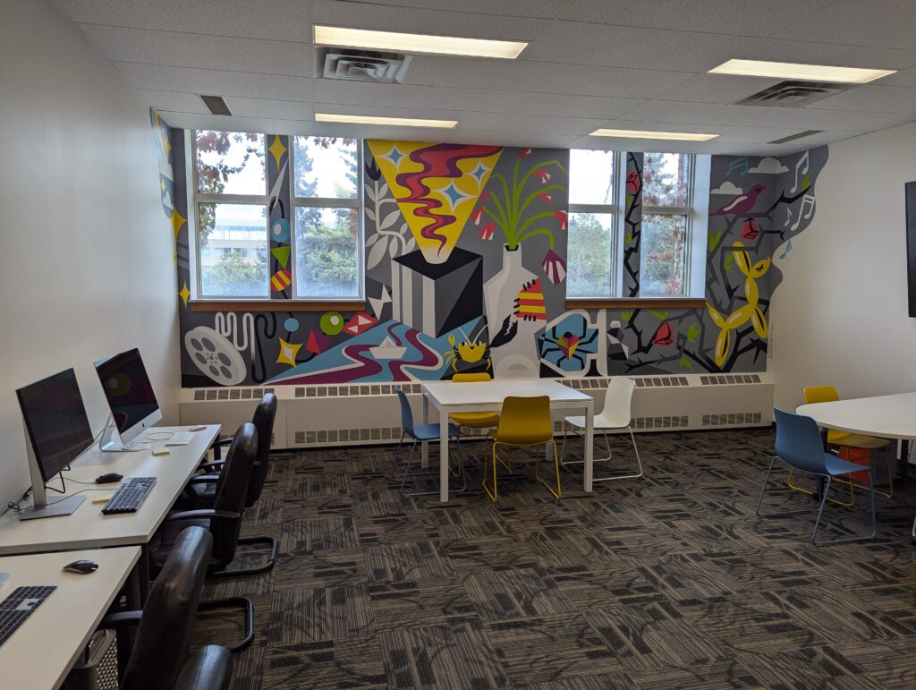 A photo of a room with tables, chairs and computers around the outside, with a large colourful mural painted on one wall.